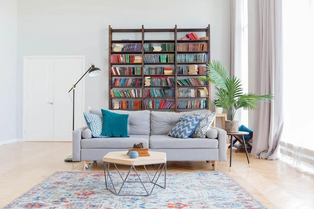 A room with standing bookcase filled with books standing behind a grey couch