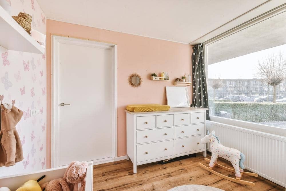 Pink wall kids room with white drawers and a large wide window