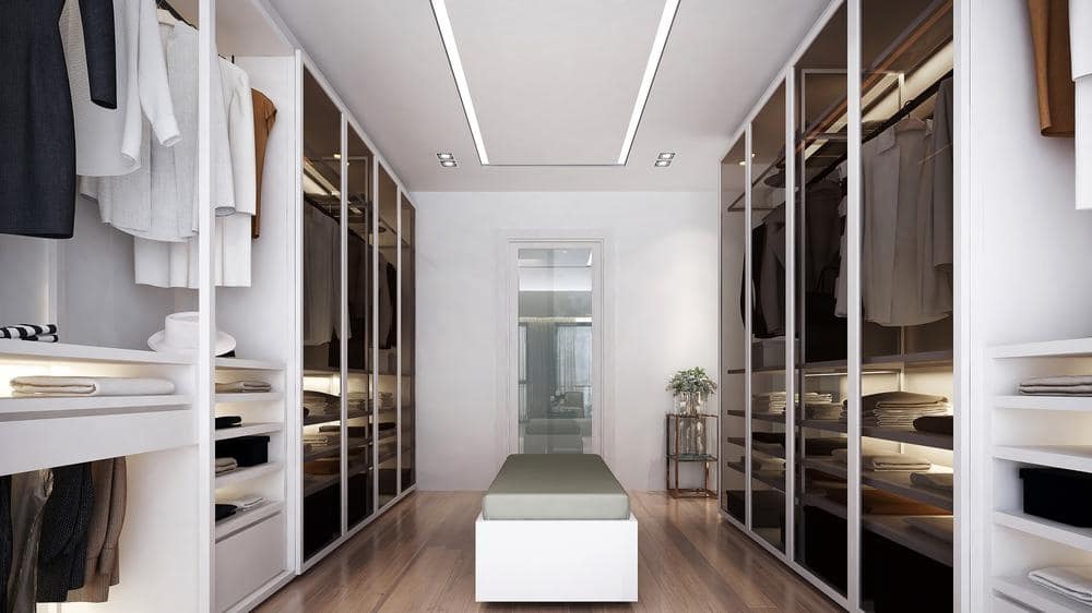 White walk in closet with dark glass doors and an ottoman